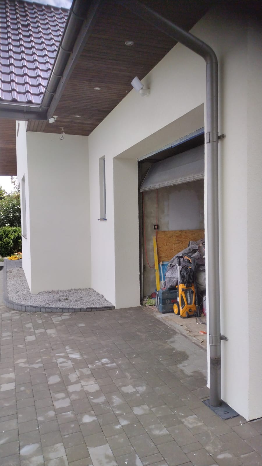 Świeżo pomalowana, jasna elewacja domu z widoczną rynną, kostką brukową i otwartym garażem z narzędziami w tle.