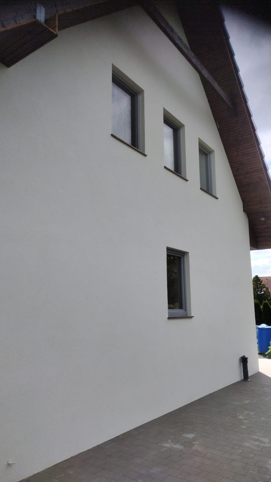 Jasna elewacja domu jednorodzinnego z czterema oknami, widok z boku, widoczna część dachu z drewnianą podbitką, szara kostka brukowa na podjeździe.