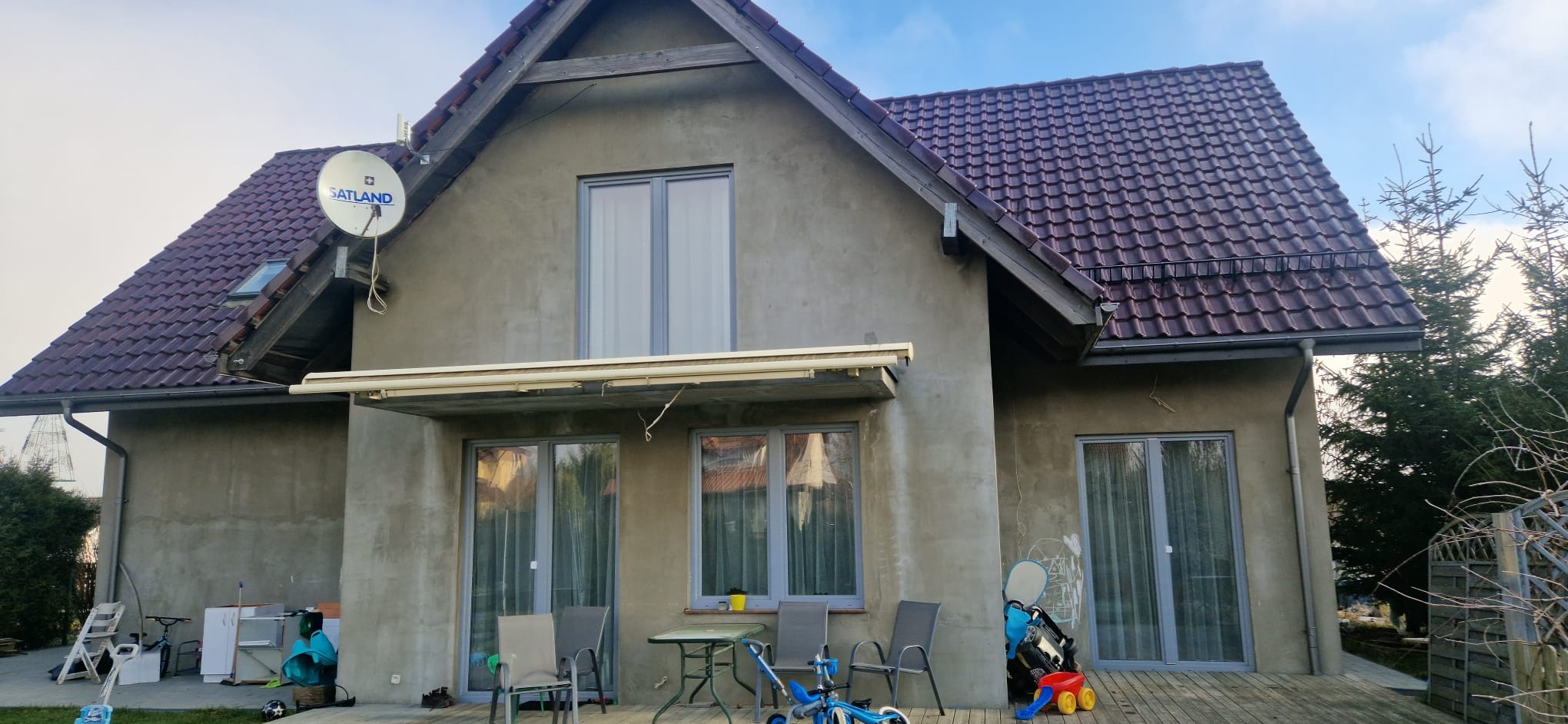 Dom jednorodzinny w trakcie prac elewacyjnych, widoczna surowa szara elewacja bez ostatecznego wykończenia, z zamontowanymi oknami i satelitą na dachu, otoczenie z elementami placu zabaw i meblami...