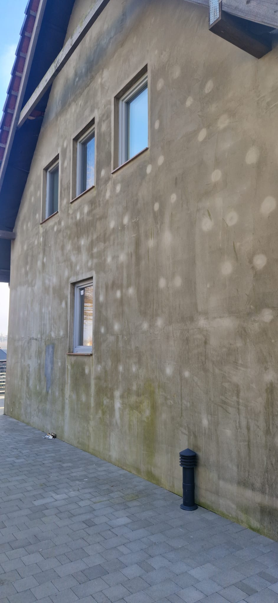 Szara elewacja budynku z widocznymi plamami i zabrudzeniami, cztery okna na różnych wysokościach, fragment dachu i chodnik z kostki brukowej u podstawy ściany.