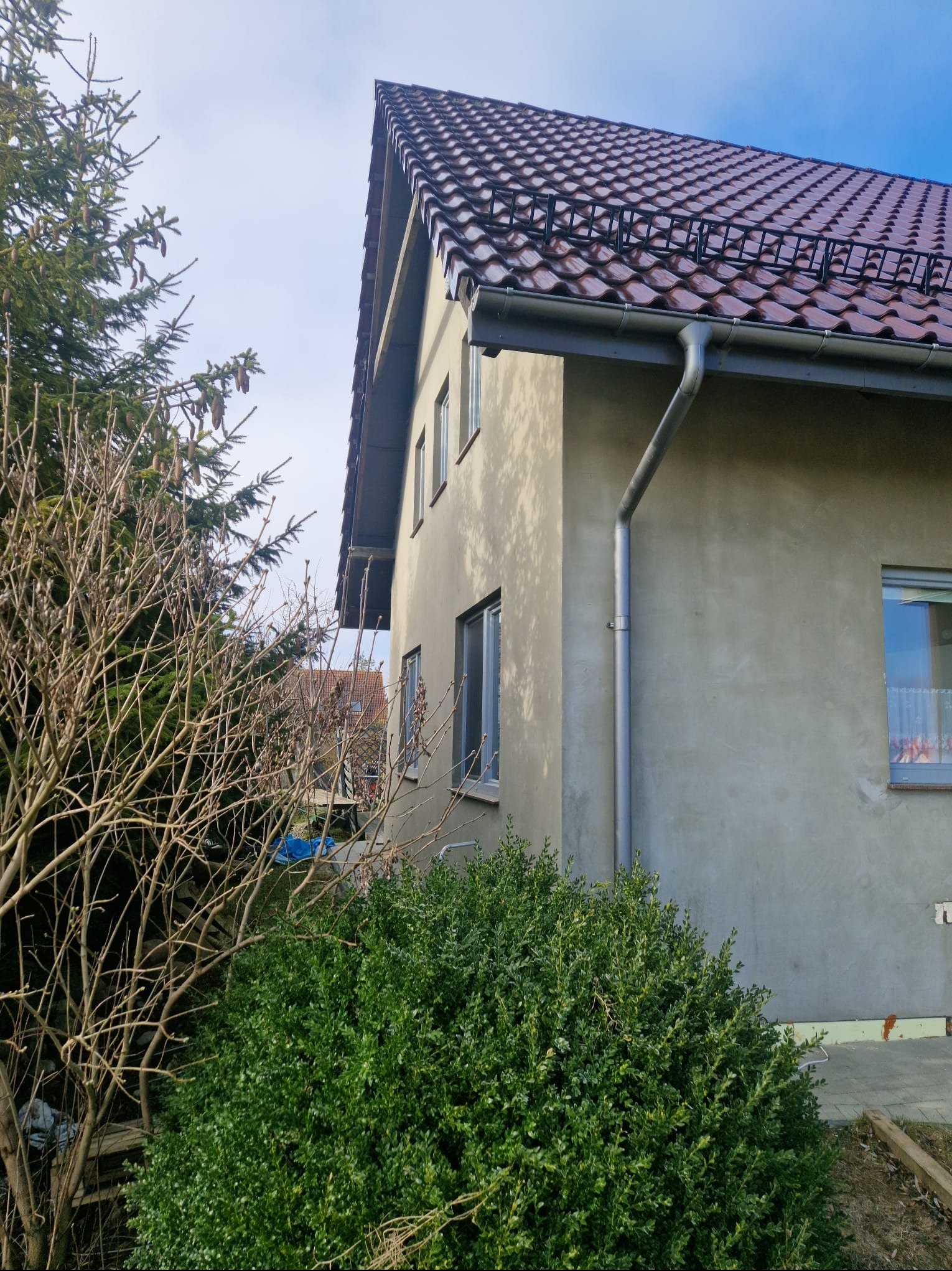 Surowa elewacja domu jednorodzinnego z widocznymi oknami, rynnami i brązowym dachem z dachówki, częściowo zasłonięta przez krzewy i drzewo iglaste bez liści, w tle widoczny fragment sąsiedniego...