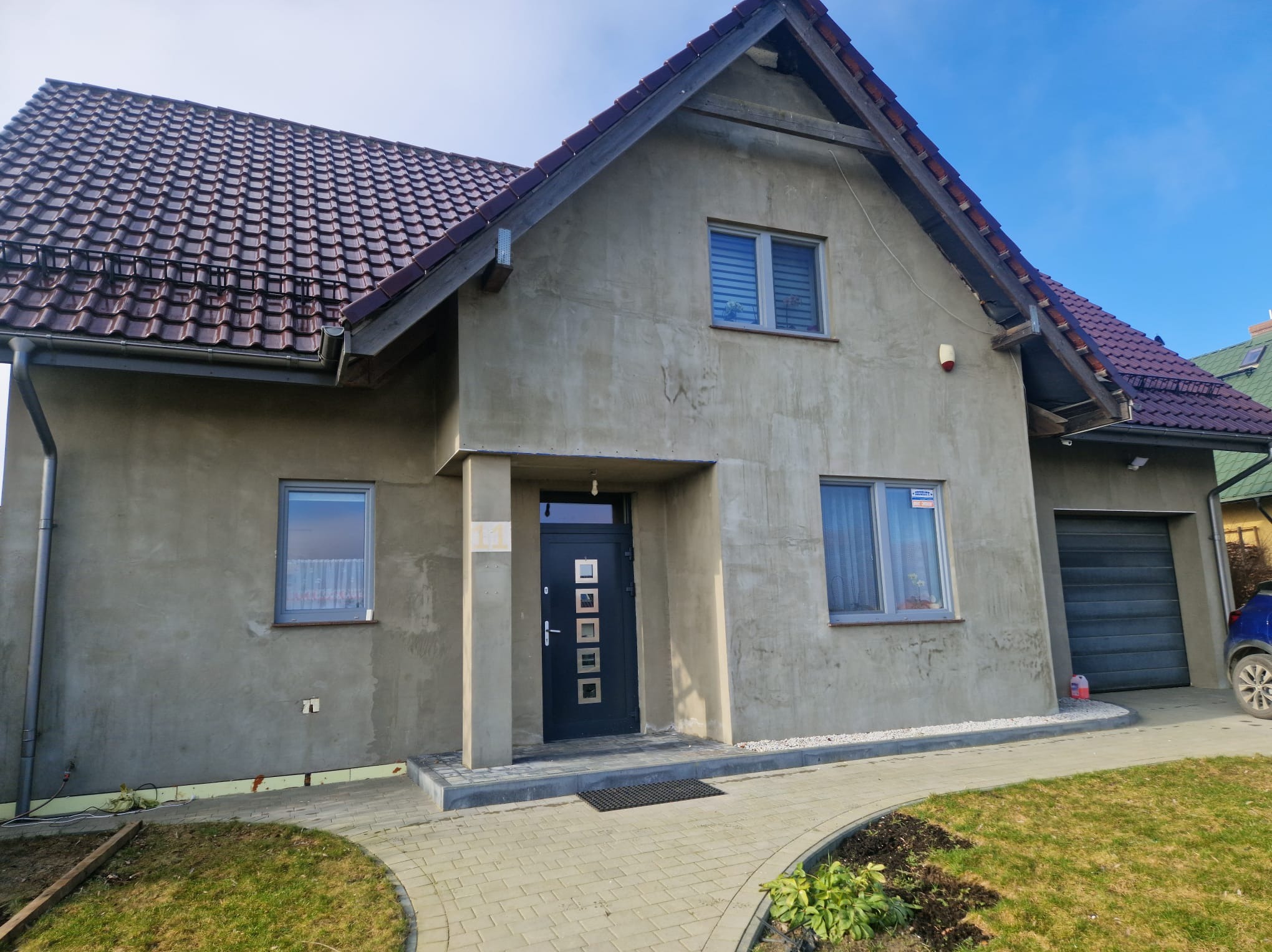 Dom jednorodzinny w trakcie prac elewacyjnych; surowy tynk szary, okna PCV z roletami, brązowa dachówka, kostka brukowa na ścieżce, widoczne fragmenty ocieplenia przy gruncie.