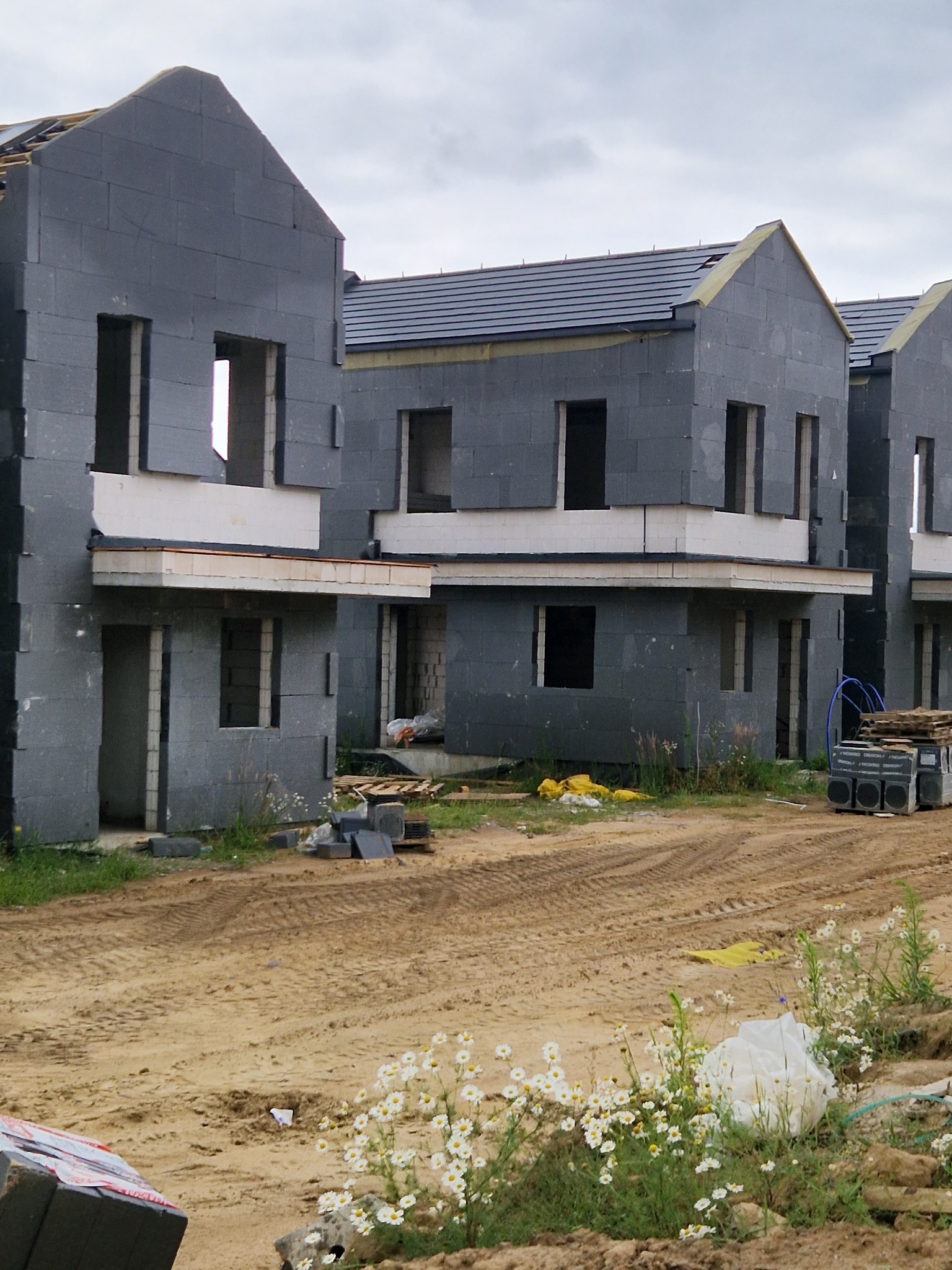 Surowe, szare domy w zabudowie szeregowej, w trakcie budowy, z widocznymi blokami elewacyjnymi i częściowo wykończonym dachem, otoczone piaszczystym terenem z polnymi kwiatami na pierwszym planie.