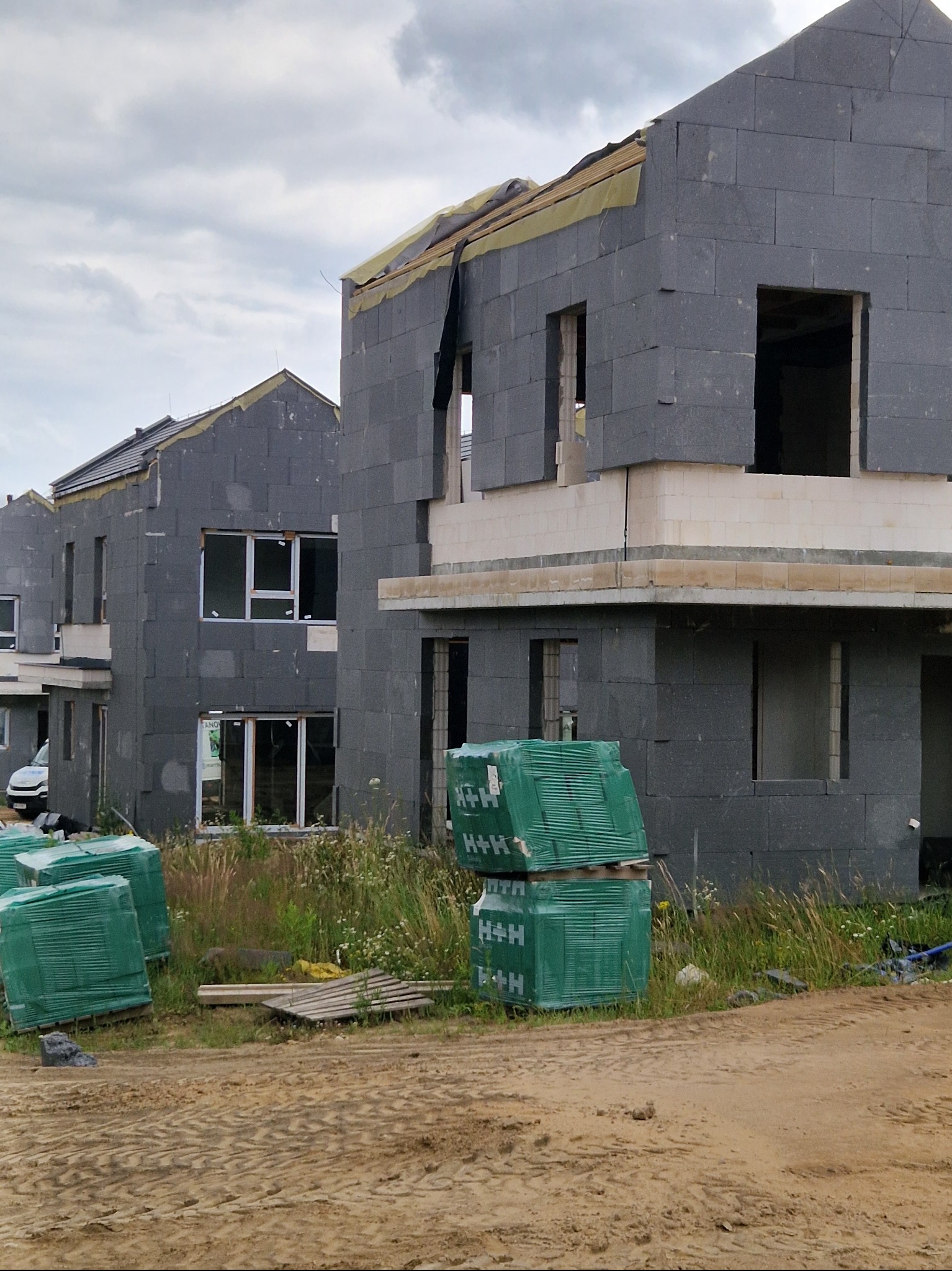 Surowe budynki z szarego bloczka, w trakcie budowy, z widocznymi paletami zielonych materiałów budowlanych H+H, na piaszczystym terenie budowy pod pochmurnym niebem.
