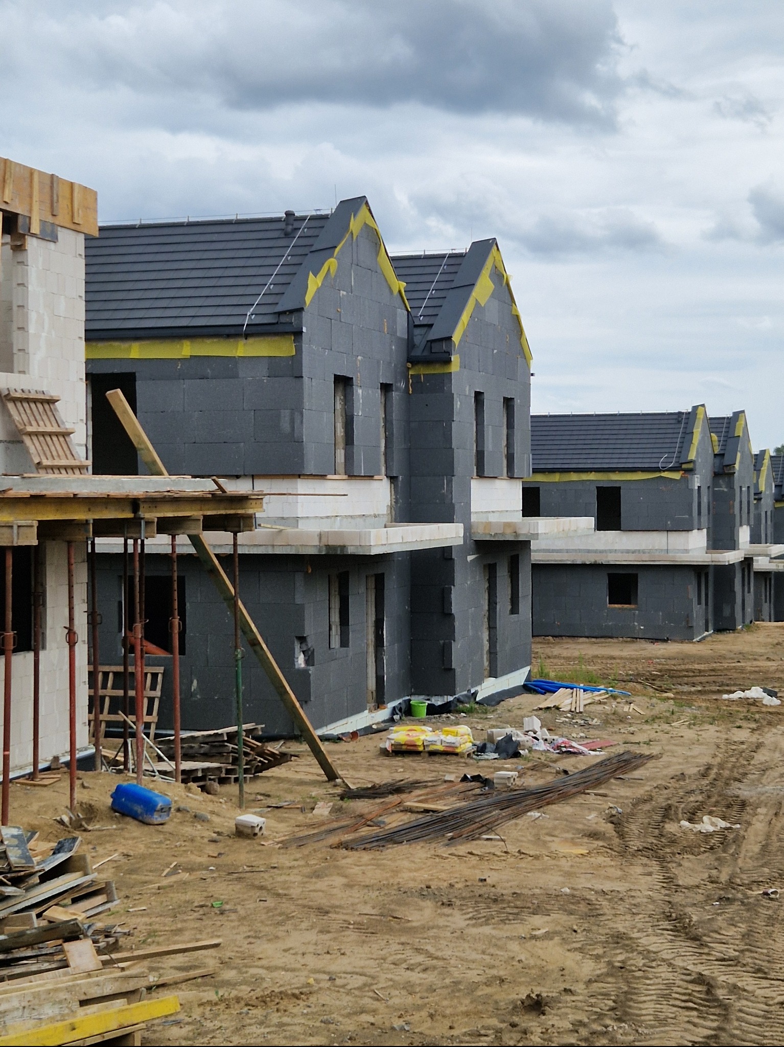 Szereg nowo budowanych domów jednorodzinnych w trakcie prac związanych z elewacją, widoczne ocieplenie ścian styropianem i przygotowanie do tynkowania, teren budowy z materiałami i narzędziami.