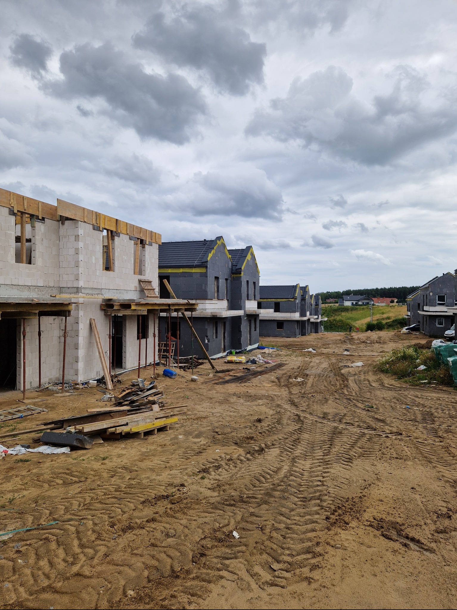 Szereg nowo budowanych domów jednorodzinnych w stanie surowym, z widoczną konstrukcją ścian z betonu komórkowego i drewnianym szalunkiem, część budynków z czarnym dachem i szarą elewacją...