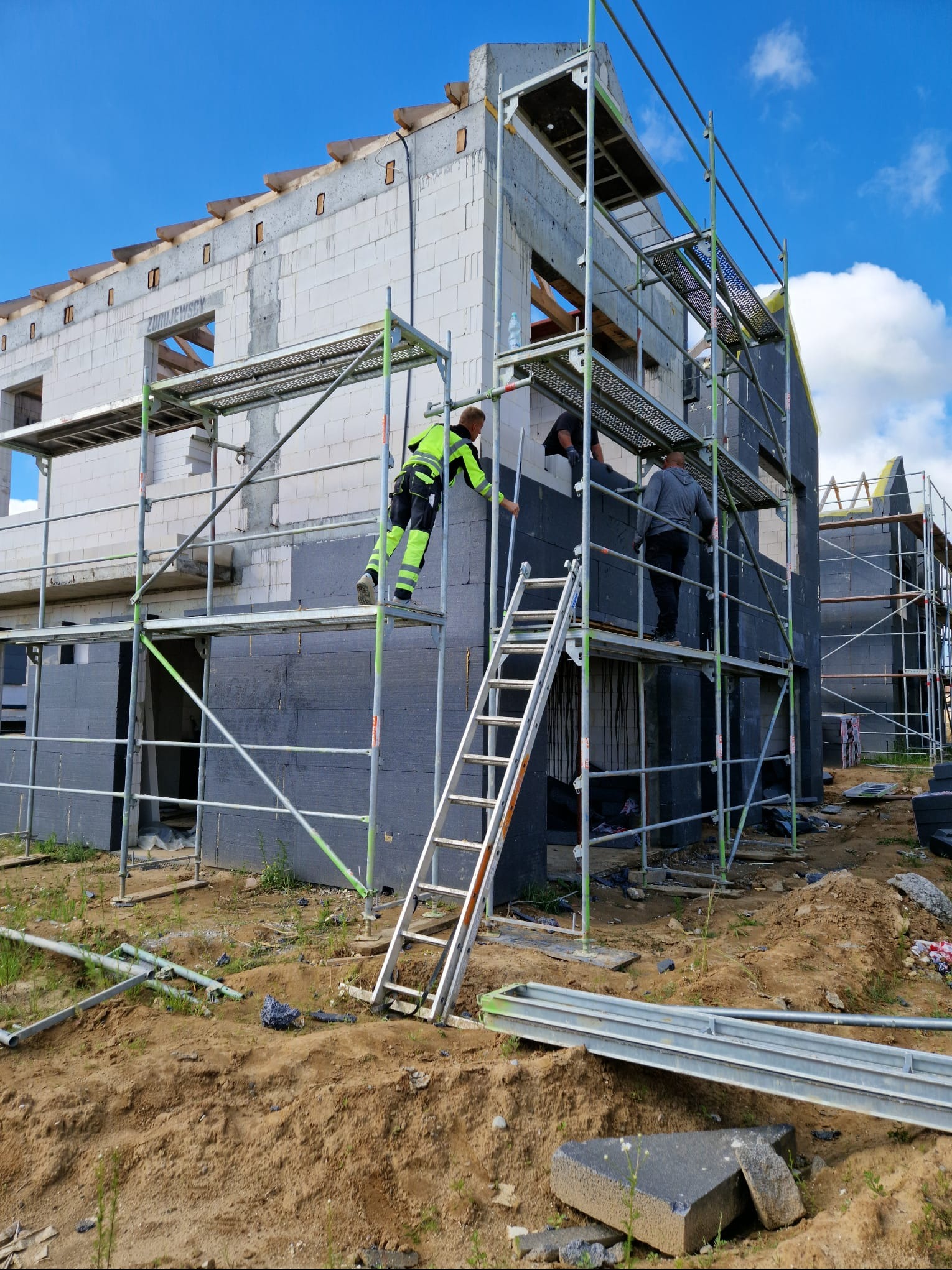 Trzech pracowników na rusztowaniu montuje grafitowe płyty izolacyjne na ścianach surowego budynku z białych bloczków, widoczne elementy konstrukcyjne dachu i błękitne niebo.