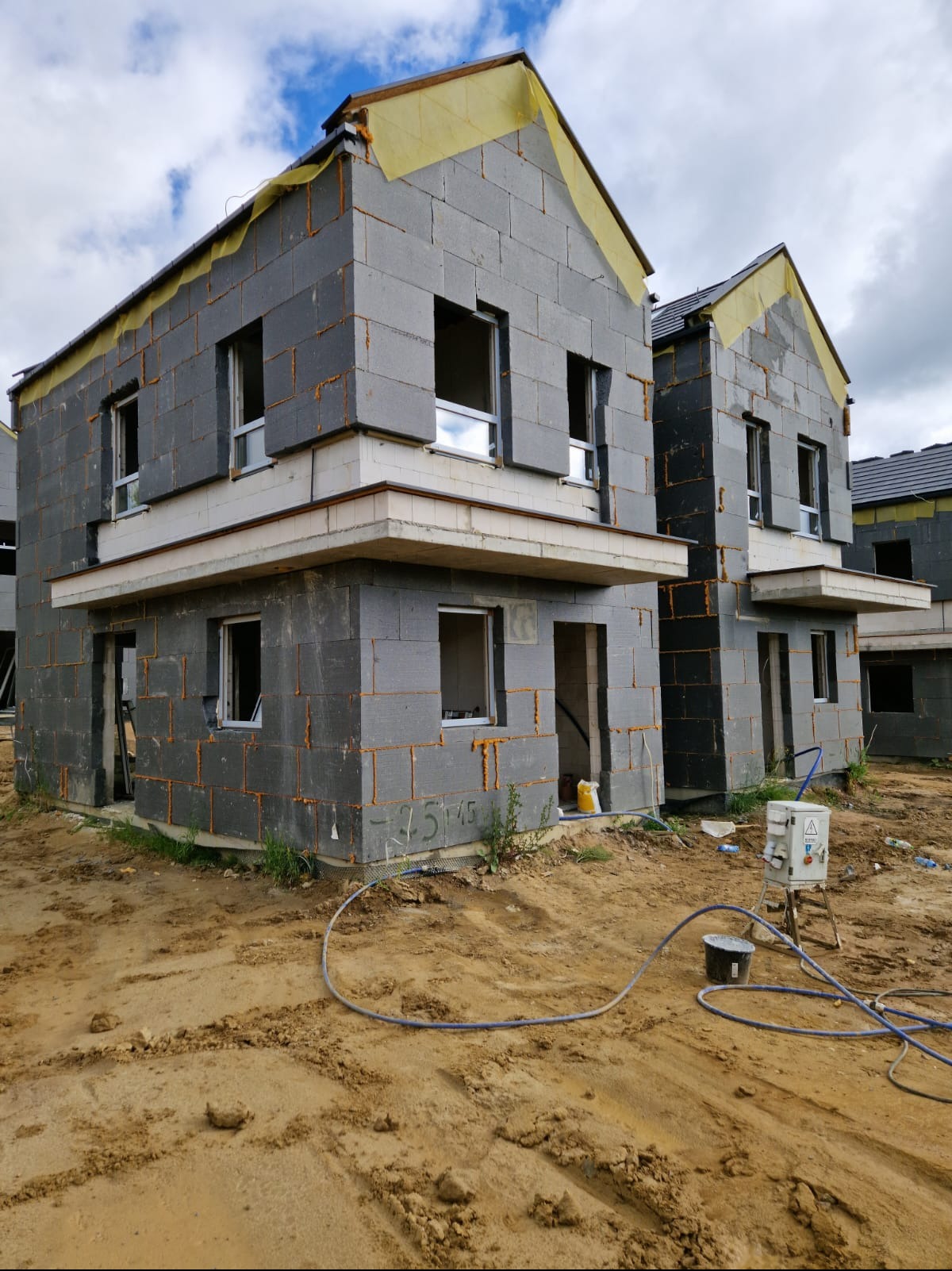 Domy w stanie surowym otwartym z szarych bloczków, widoczne przygotowania do dalszych prac budowlanych, teren wokół niezagospodarowany, z widocznymi śladami budowy i piaskiem.