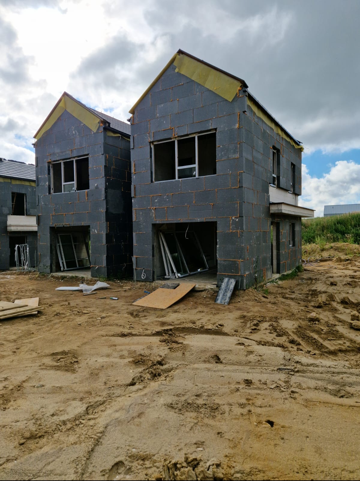 Surowe domy w zabudowie szeregowej, w trakcie wznoszenia ścian z bloczków, z widocznym ociepleniem na szczytach, teren budowy z piaskiem i porozrzucanymi materiałami.