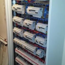 Rozdzielnica elektryczna z zainstalowanymi bezpiecznikami, stycznikami i okablowaniem, widoczne przewody w kolorach niebieskim, czerwonym i żółto-zielonym, częściowo zasłonięta obudową w kolorze...