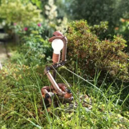 Oryginalna lampa ogrodowa z rur w stylu steampunk, z żarówką i kapeluszem, umieszczona w zieleni ogrodu w Zaczerniu.