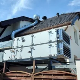 Zewnętrzna instalacja rekuperacji z metalowymi kanałami wentylacyjnymi na dachu budynku jednorodzinnego, widok z poziomu ogrodzenia.