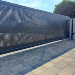 Nowoczesna, przesuwna brama wjazdowa z perforowanej blachy w kolorze antracytowym, z widocznymi cieniami na kostce brukowej. Tło stanowi betonowe ogrodzenie i zieleń.