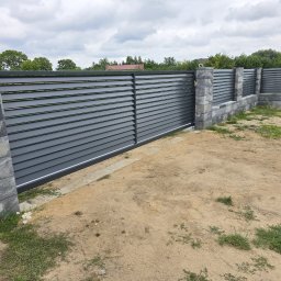 OLI LOOK Usługi Ślusarskie Hubert Podlesiński - Nowoczesna, przesuwna brama wjazdowa z poziomych, szarych paneli, osadzona na kamiennych słupkach. Tło: zielona roślinność i pochmurne niebo.