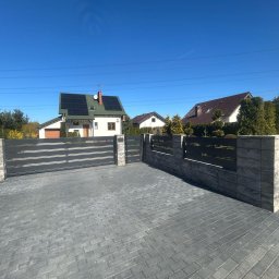 OLI LOOK Usługi Ślusarskie Hubert Podlesiński - Nowoczesna brama wjazdowa z szarego metalu i kamiennymi słupkami, otoczona kostką brukową. W tle domy z panelami słonecznymi i błękitne niebo, tworzące spójny, estetyczny wygląd.