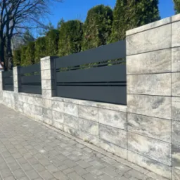 Nowoczesne ogrodzenie z szarej, poziomej blachy i kamiennych słupków w Golina. Widok na chodnik i drzewa w tle. Minimalistyczny design.