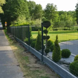 Nowoczesne ogrodzenie panelowe w kolorze grafitowym z podmurówką betonową, obsadzone ozdobnymi krzewami, widok od strony ulicy z zielenią w tle.