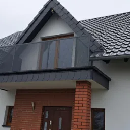 Dom jednorodzinny z balkonem zabezpieczonym nowoczesną, ażurową balustradą w kolorze antracytowym, dopasowaną do koloru dachu i orynnowania. Widoczne ceglane filary podtrzymujące balkon.