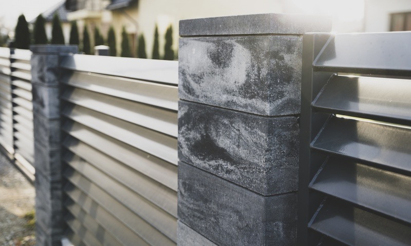 Nowoczesne ogrodzenie panelowe z betonowymi słupkami. Metalowe przęsła w szarym kolorze, ułożone poziomo, tworzą minimalistyczny design. Widoczne detale łączeń i faktura betonu.