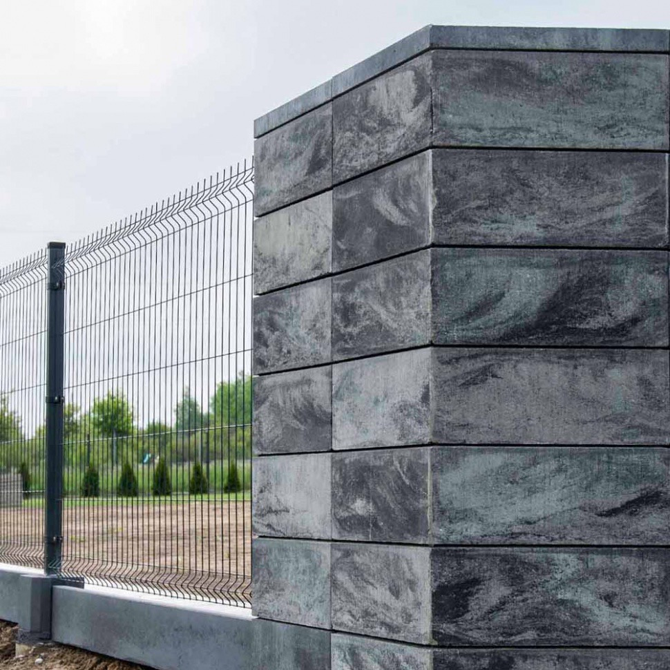 Nowoczesne ogrodzenie panelowe z betonowym słupkiem o marmurkowej fakturze. Szara kolorystyka, siatka metalowa. Widok z bliska na tle zieleni.