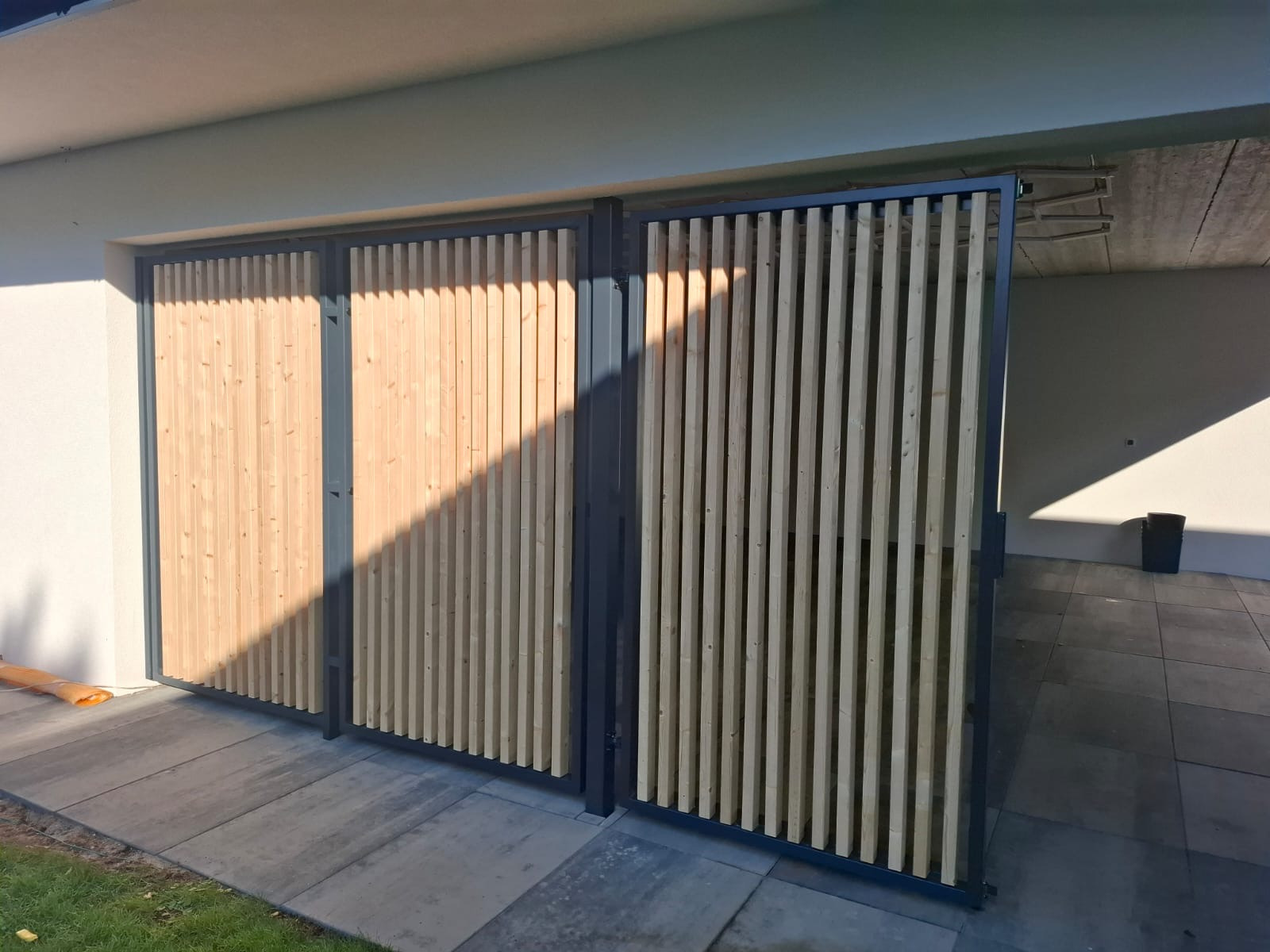 Nowoczesna brama wjazdowa z drewnianymi lamelami w szarej metalowej ramie, z delikatnym cieniem na jasnym tle elewacji.