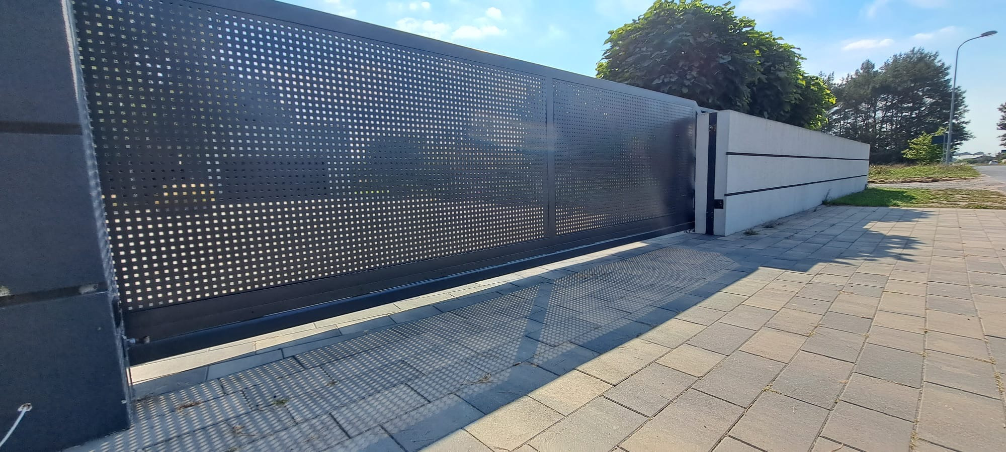Nowoczesna, przesuwna brama wjazdowa z perforowanej blachy w kolorze antracytowym, z widocznymi cieniami na kostce brukowej. Tło stanowi betonowe ogrodzenie i zieleń.