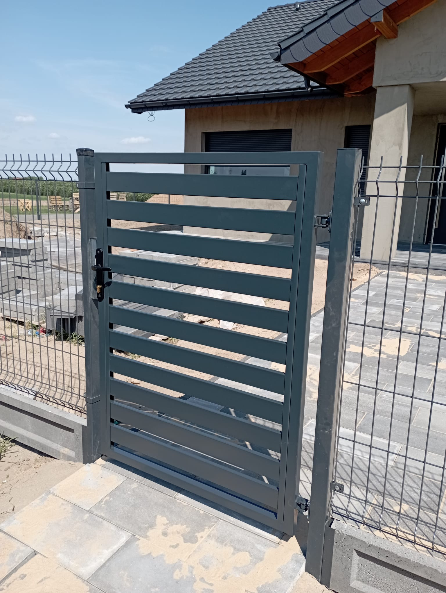 Nowoczesna, antracytowa brama wjazdowa z poziomymi lamelami, z czarną klamką, dopasowana do ogrodzenia panelowego na tle domu z szarą dachówką.