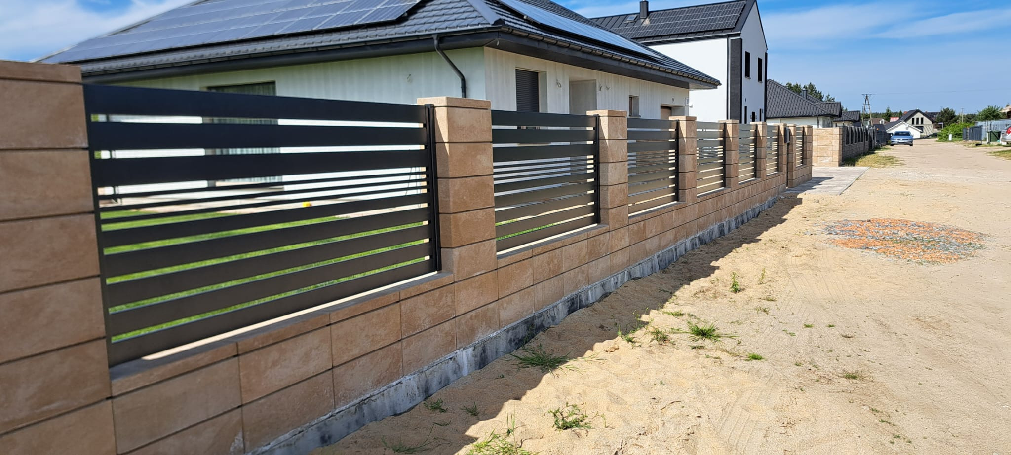 Nowoczesne ogrodzenie panelowe z poziomymi, czarnymi przęsłami i słupkami z beżowych bloczków, na tle domu z panelami słonecznymi.