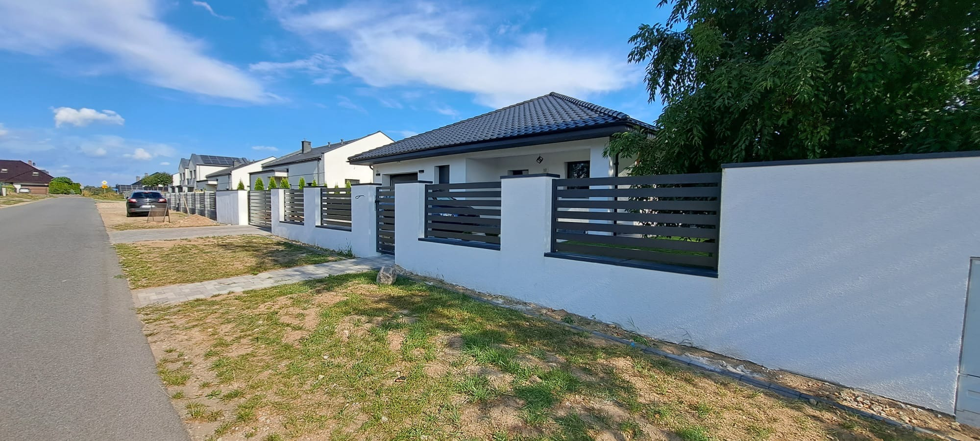 Nowoczesne ogrodzenie panelowe z poziomymi, grafitowymi przęsłami i białymi słupkami, otaczające dom z ciemną dachówką na tle błękitnego nieba.