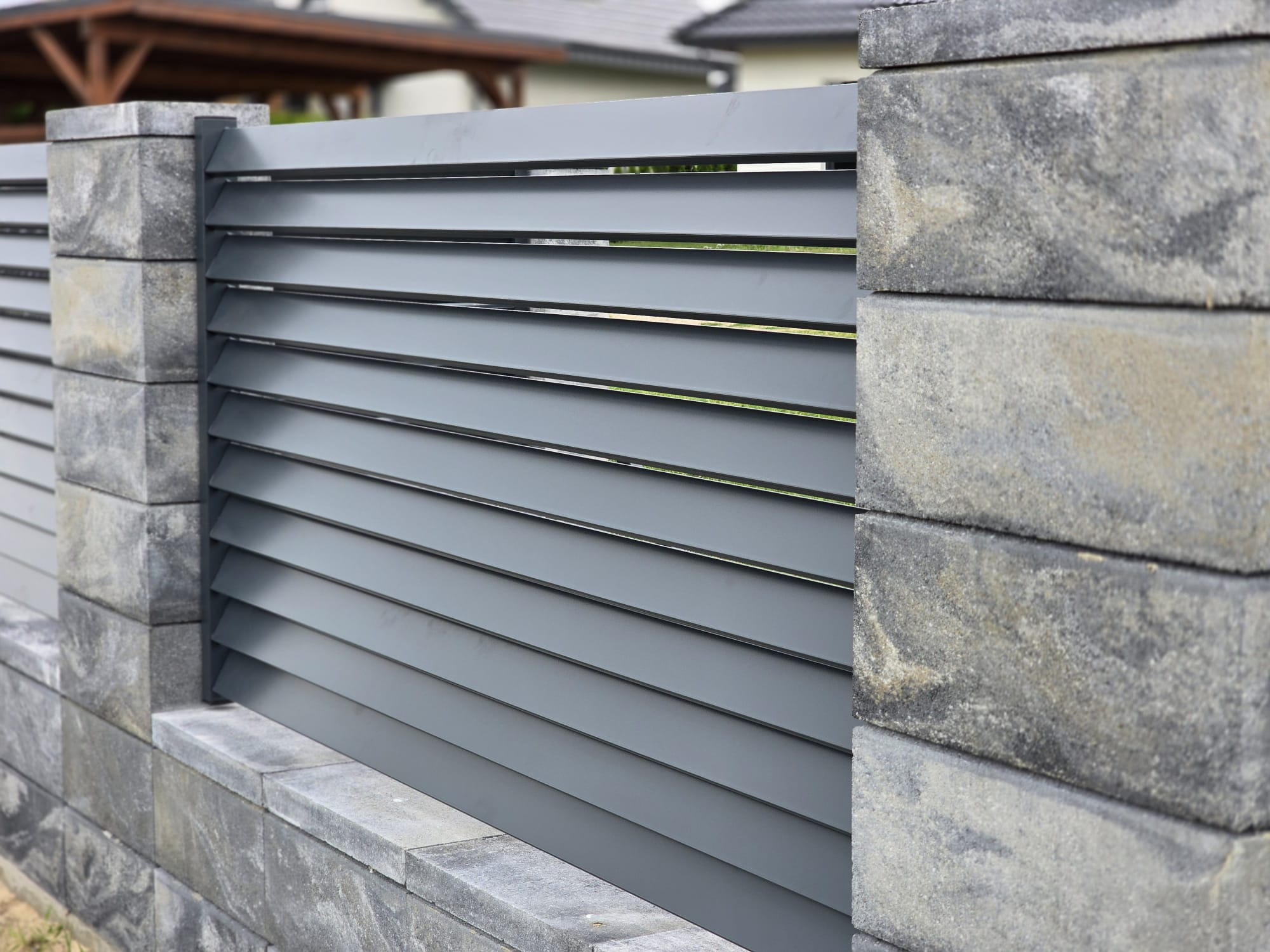 Nowoczesne ogrodzenie panelowe z szarego metalu i kamiennych słupków. Widoczne poziome lamele i faktura kamienia. Tło z zielenią i fragmentami zabudowy.