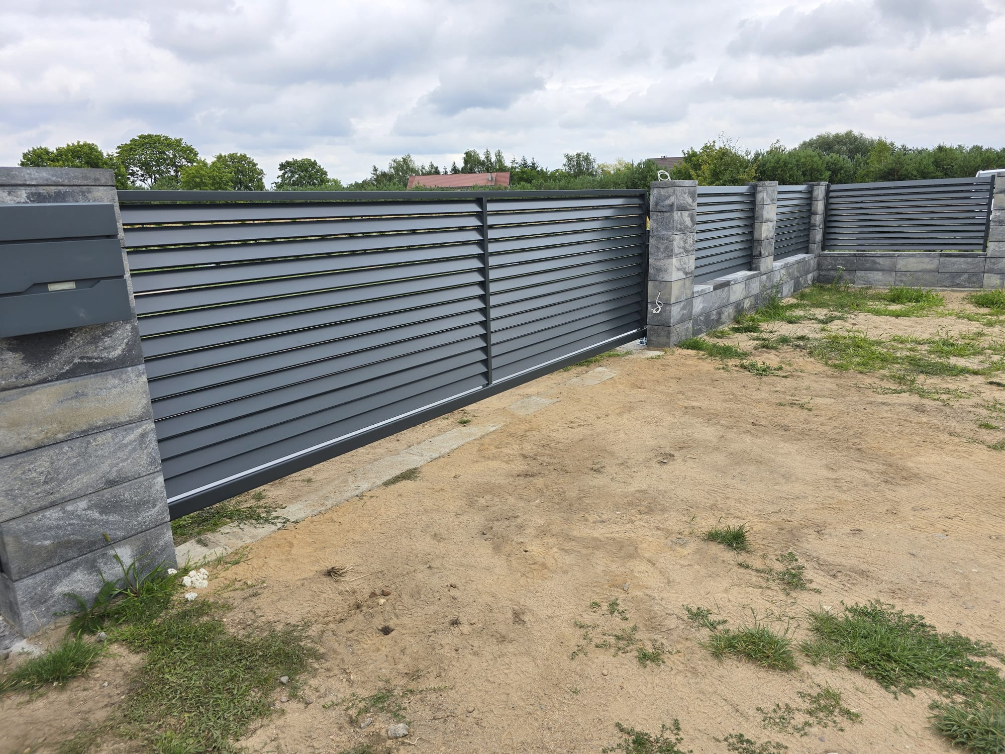 Nowoczesna, przesuwna brama wjazdowa z poziomych, szarych paneli, osadzona na kamiennych słupkach. Tło: zielona roślinność i pochmurne niebo.
