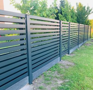 Nowoczesne, antracytowe ogrodzenie panelowe z poziomymi lamelami na betonowej podmurówce, otoczone zielonym trawnikiem i roślinnością.