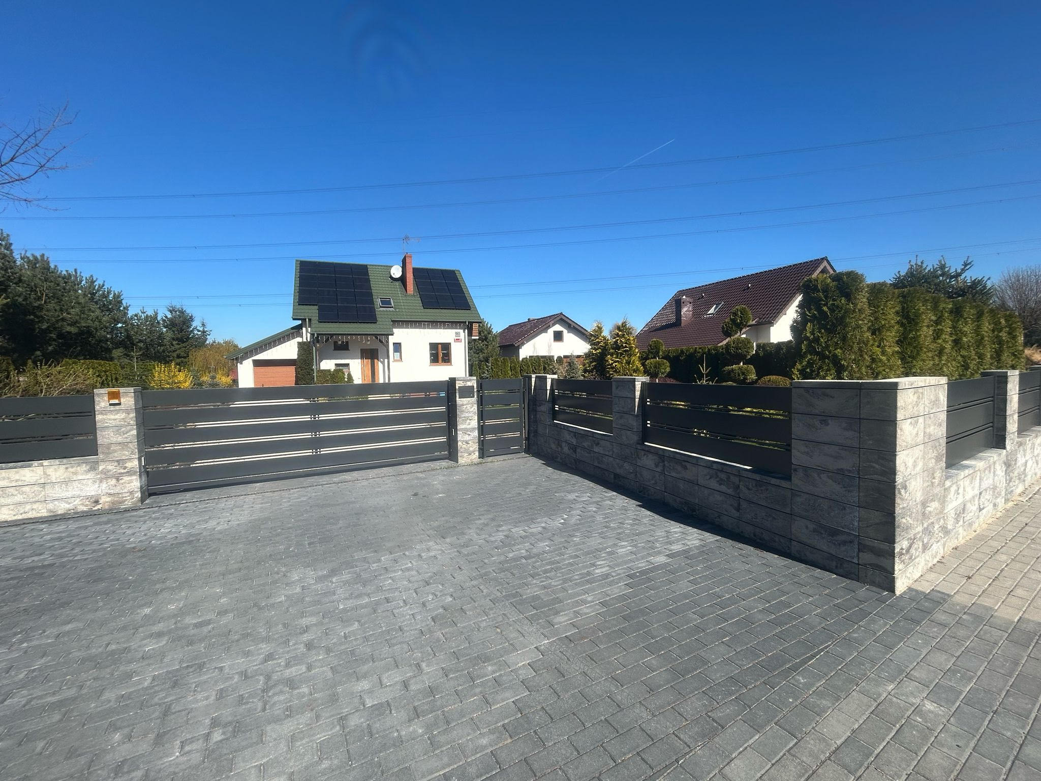 Nowoczesna brama wjazdowa z szarego metalu i kamiennymi słupkami, otoczona kostką brukową. W tle domy z panelami słonecznymi i błękitne niebo, tworzące spójny, estetyczny wygląd.