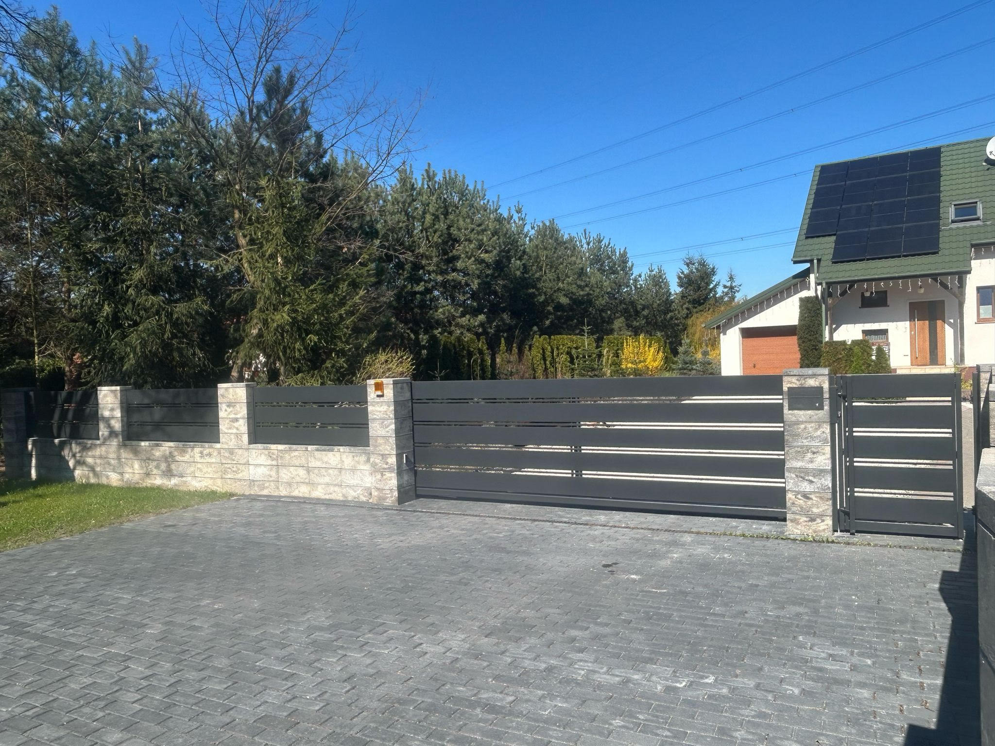 Nowoczesna brama wjazdowa z poziomymi, metalowymi przęsłami w kolorze grafitu, osadzona na murowanych słupkach z jasnego kamienia, na tle domu z panelami solarnymi.