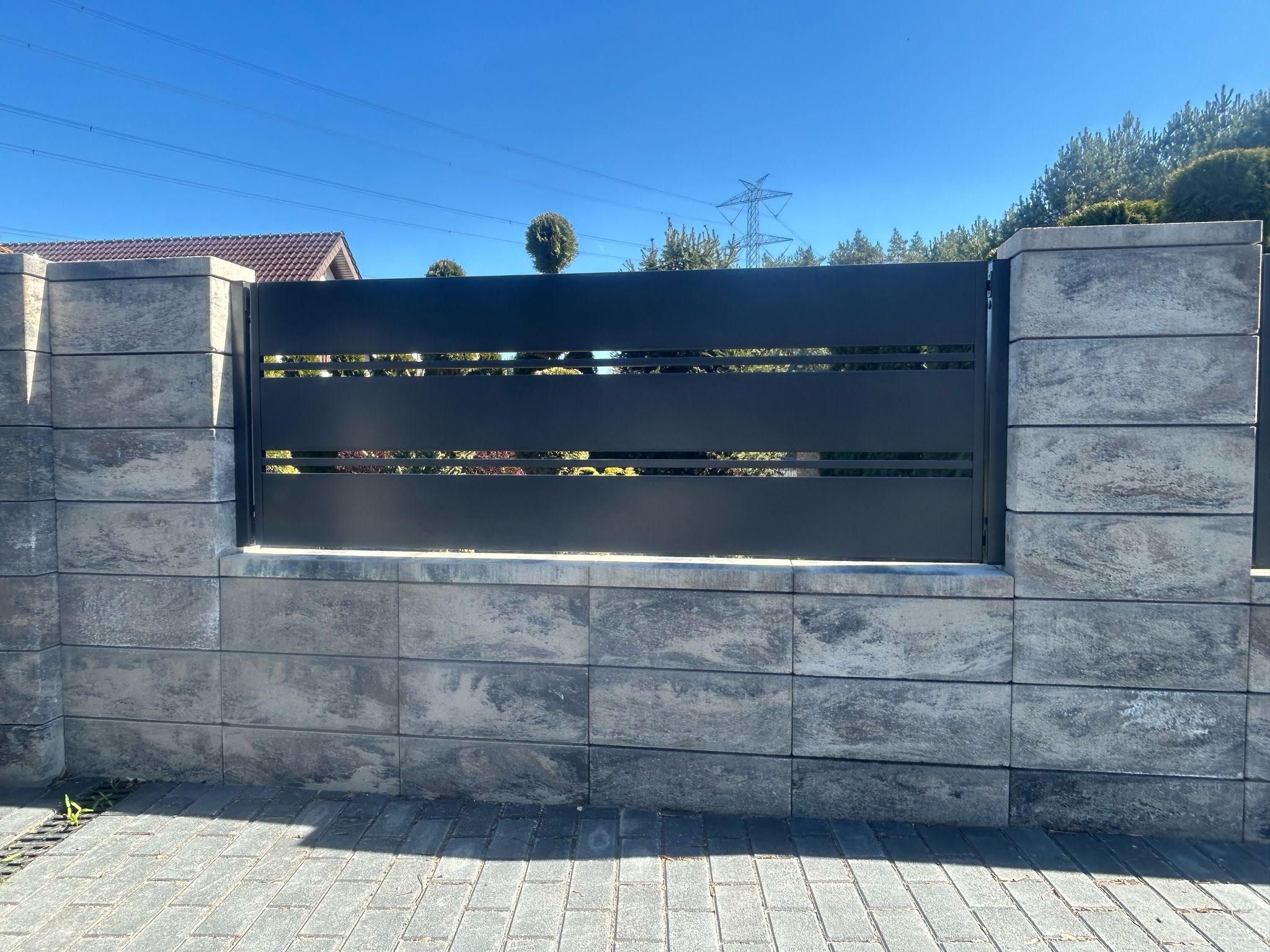 Nowoczesne, grafitowe przęsło ogrodzeniowe z poziomymi szczelinami, osadzone na szarych, betonowych słupkach. W tle błękitne niebo i linia energetyczna.