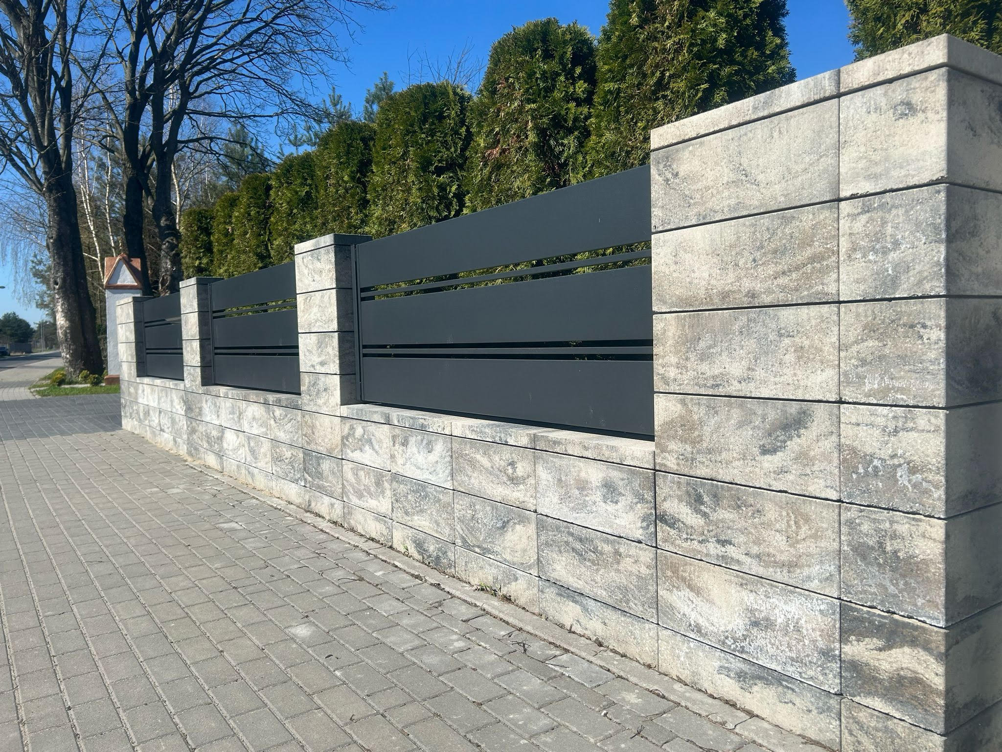 Nowoczesne ogrodzenie z szarej, poziomej blachy i kamiennych słupków w Golina. Widok na chodnik i drzewa w tle. Minimalistyczny design.