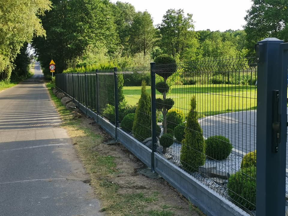 Nowoczesne ogrodzenie panelowe w kolorze grafitowym z podmurówką betonową, obsadzone ozdobnymi krzewami, widok od strony ulicy z zielenią w tle.