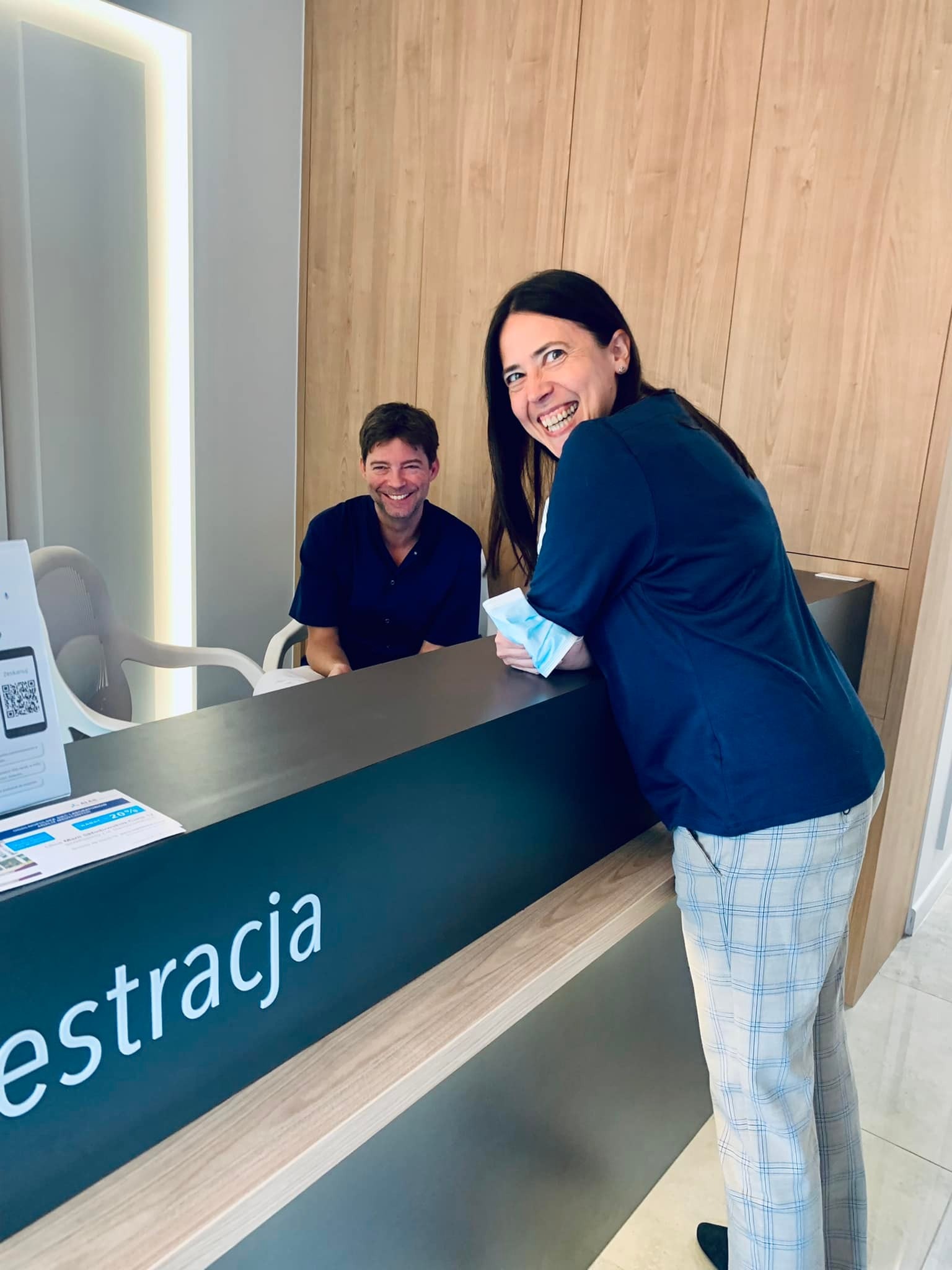 Uśmiechnięta kobieta opiera się o ladę recepcji z napisem 'rejestracja', w tle siedzący mężczyzna w gabinecie.