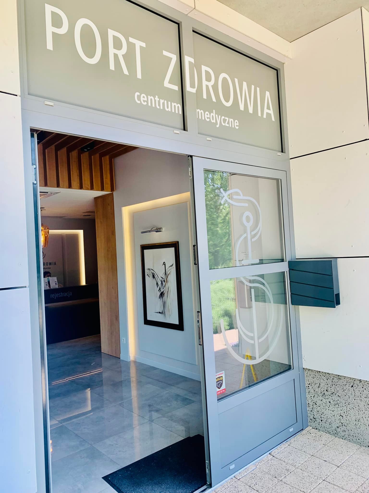 Wejście do centrum medycznego 'Port Zdrowia' z logo na szklanych drzwiach, widok na recepcję i obraz na ścianie.