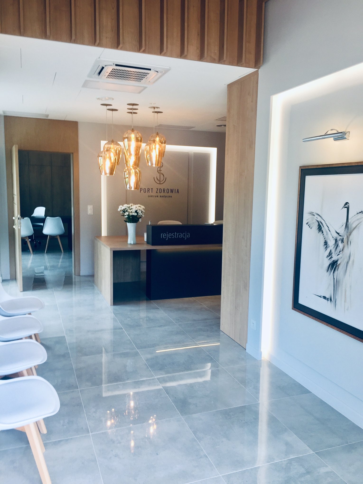 Nowoczesna poczekalnia w centrum medycznym z recepcją, krzesłami dla pacjentów, dekoracyjnymi lampami i obrazem ptaka na ścianie.