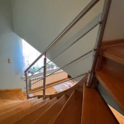 Drewniane schody spiralne z metalową balustradą, widok z góry. Na parapecie okna widoczne rośliny w doniczkach. Schody w odcieniach brązu, ściany jasne.