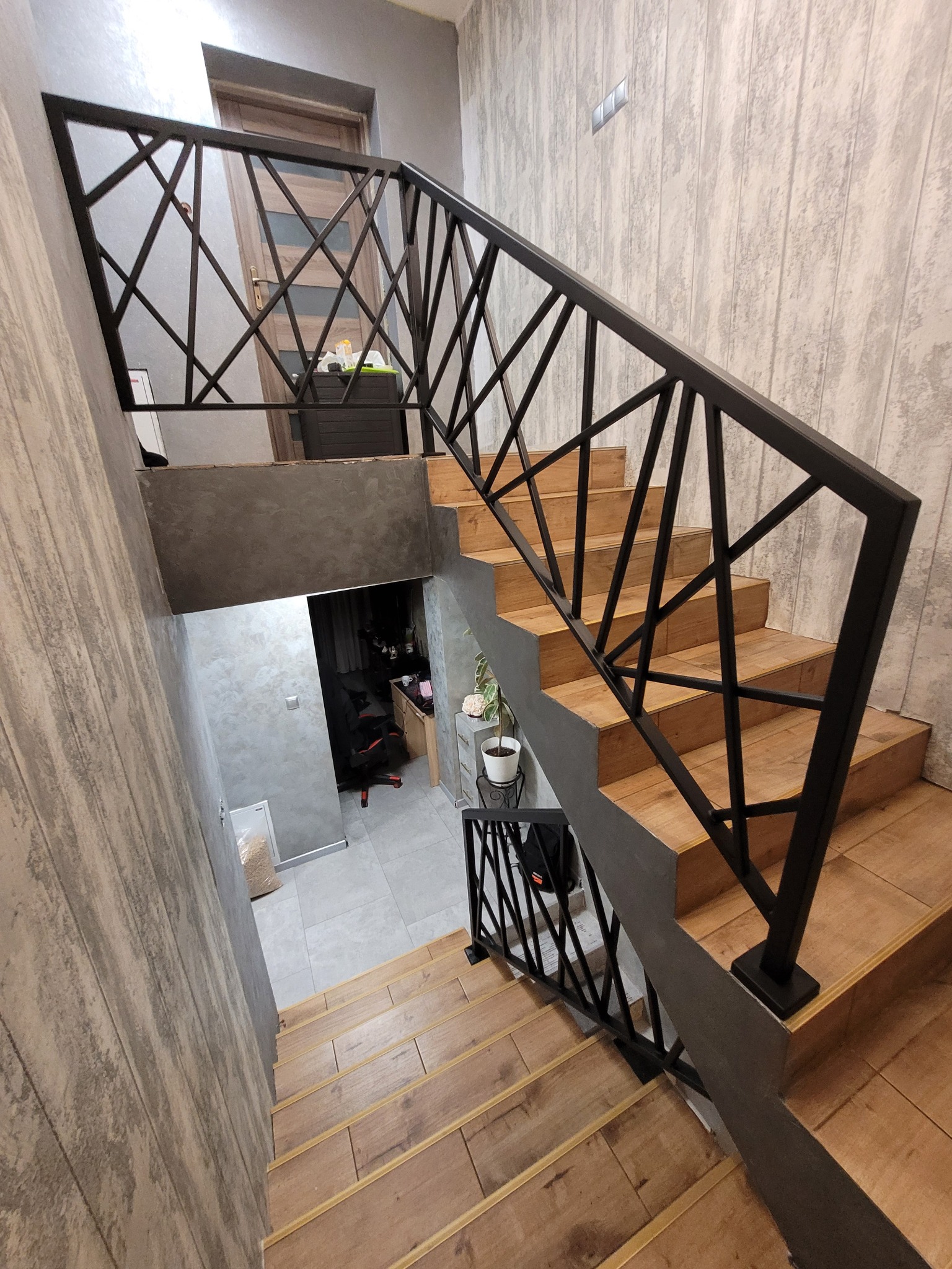 Schody drewniane z czarną, geometryczną balustradą metalową, widok z góry, szare ściany z fakturą drewna, drzwi w tle.
