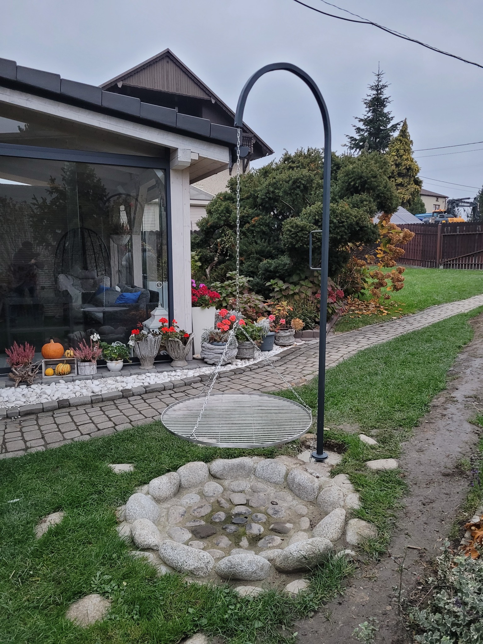 Metalowy grill wiszący nad kamiennym paleniskiem w ogrodzie, widok na przeszkloną werandę z dekoracjami jesiennymi.