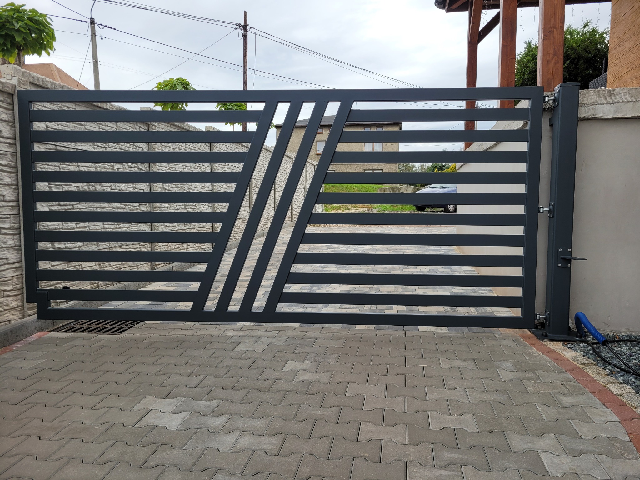 Nowoczesna, pozioma brama wjazdowa w kolorze grafitowym z ukośnymi elementami dekoracyjnymi, zamontowana na posesji z kostką brukową, widok z zewnątrz.