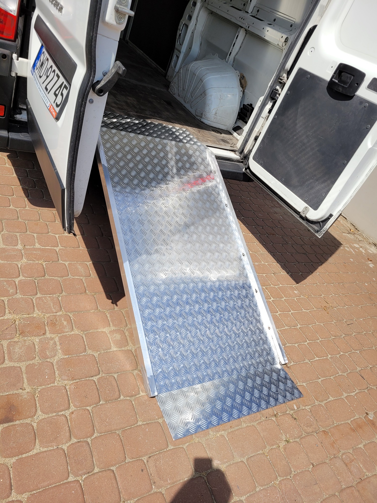 Aluminiowa rampa najazdowa zamontowana do tylnych drzwi białego samochodu dostawczego, ułatwiająca załadunek i rozładunek na brukowanym podłożu.