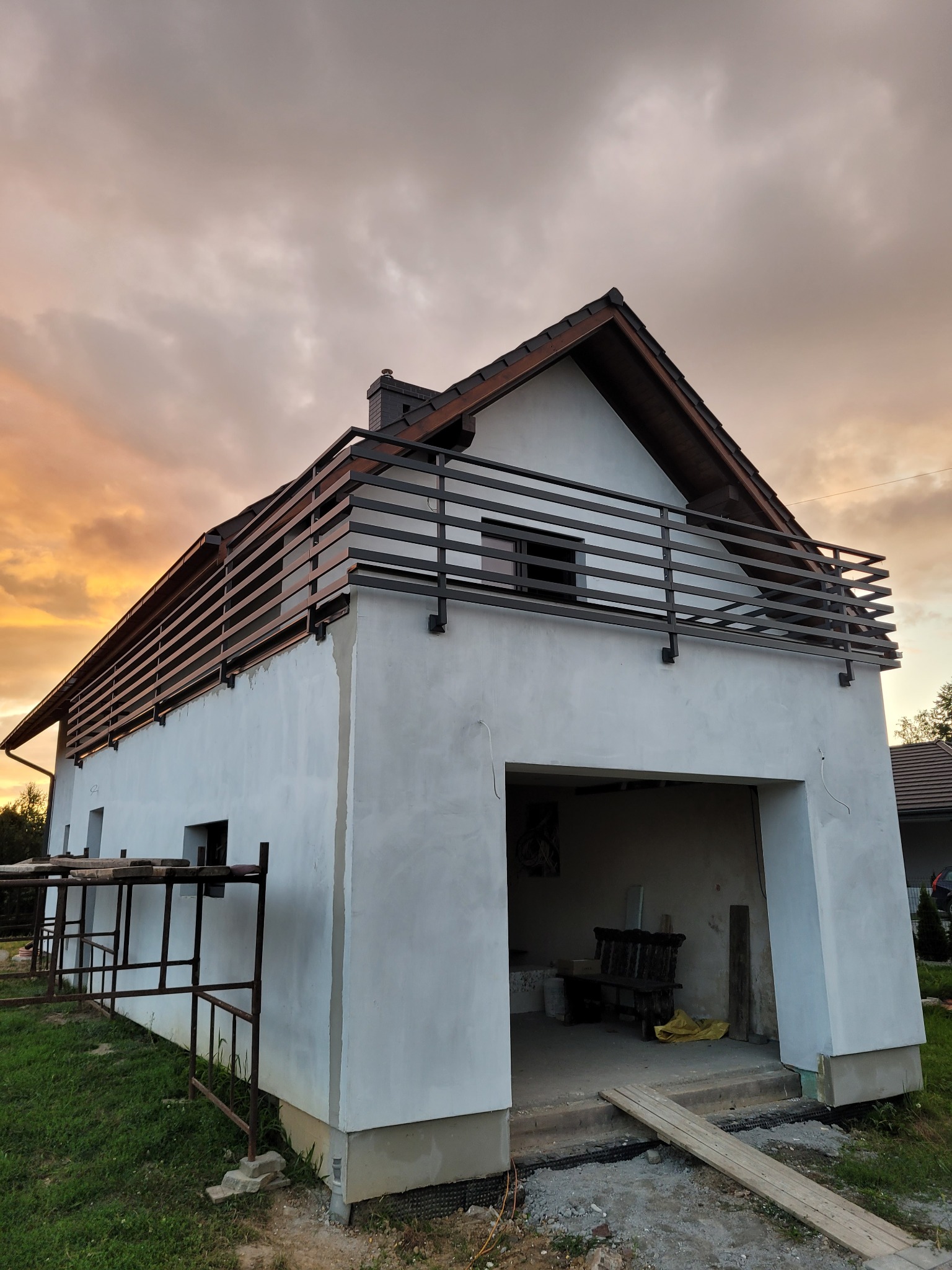 Dwukondygnacyjny dom w trakcie wykończenia z metalową balustradą balkonową na drugim piętrze, widok z dołu na tle zachodzącego słońca i częściowo zachmurzonego nieba.