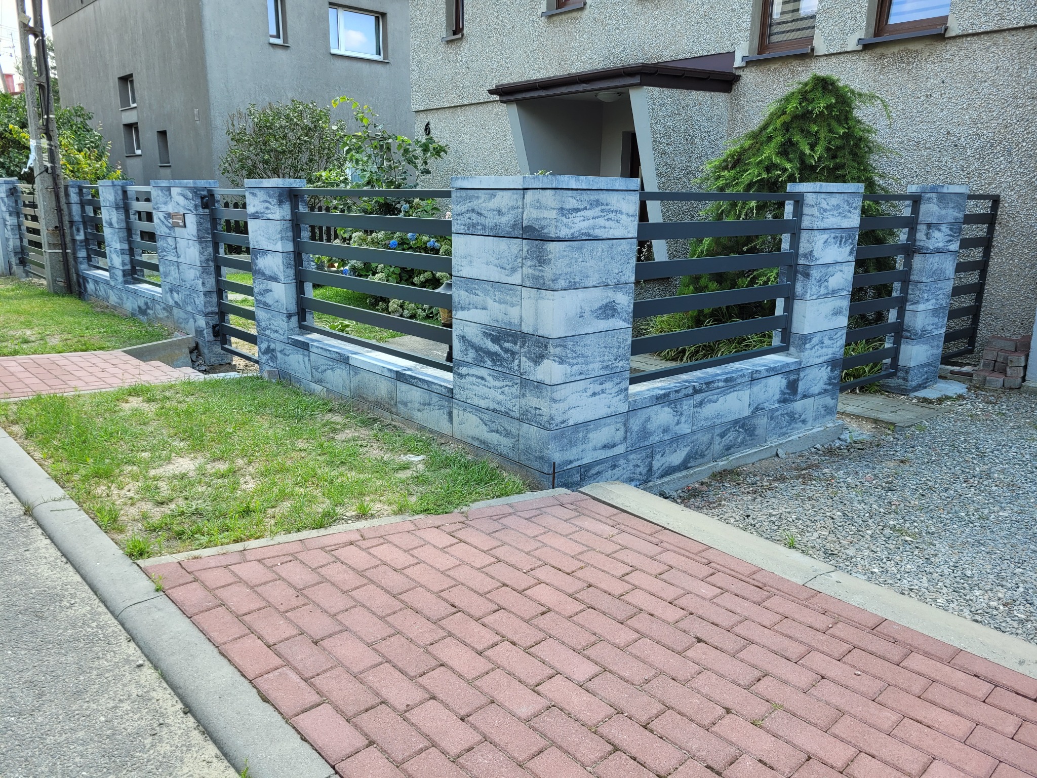 Nowoczesne ogrodzenie z szarego kamienia łupanego i poziomych, ciemnoszarych metalowych przęseł, widoczne od strony chodnika z czerwonej kostki brukowej, z budynkiem w tle.