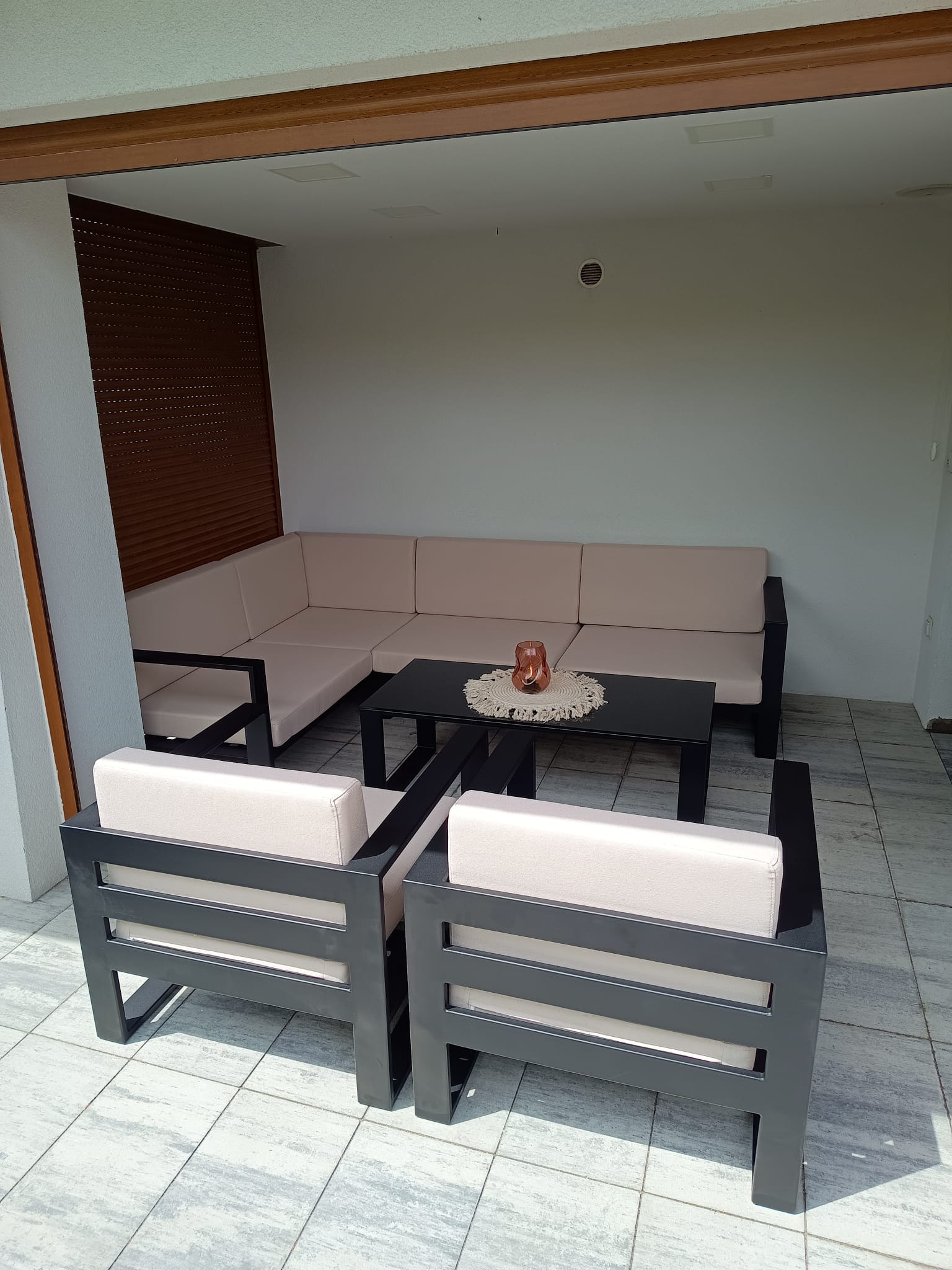 Zadaszony taras z szarymi metalowymi meblami ogrodowymi: narożna sofa z jasnymi poduszkami, dwa fotele i czarny stolik kawowy z ozdobną serwetką i świecą.