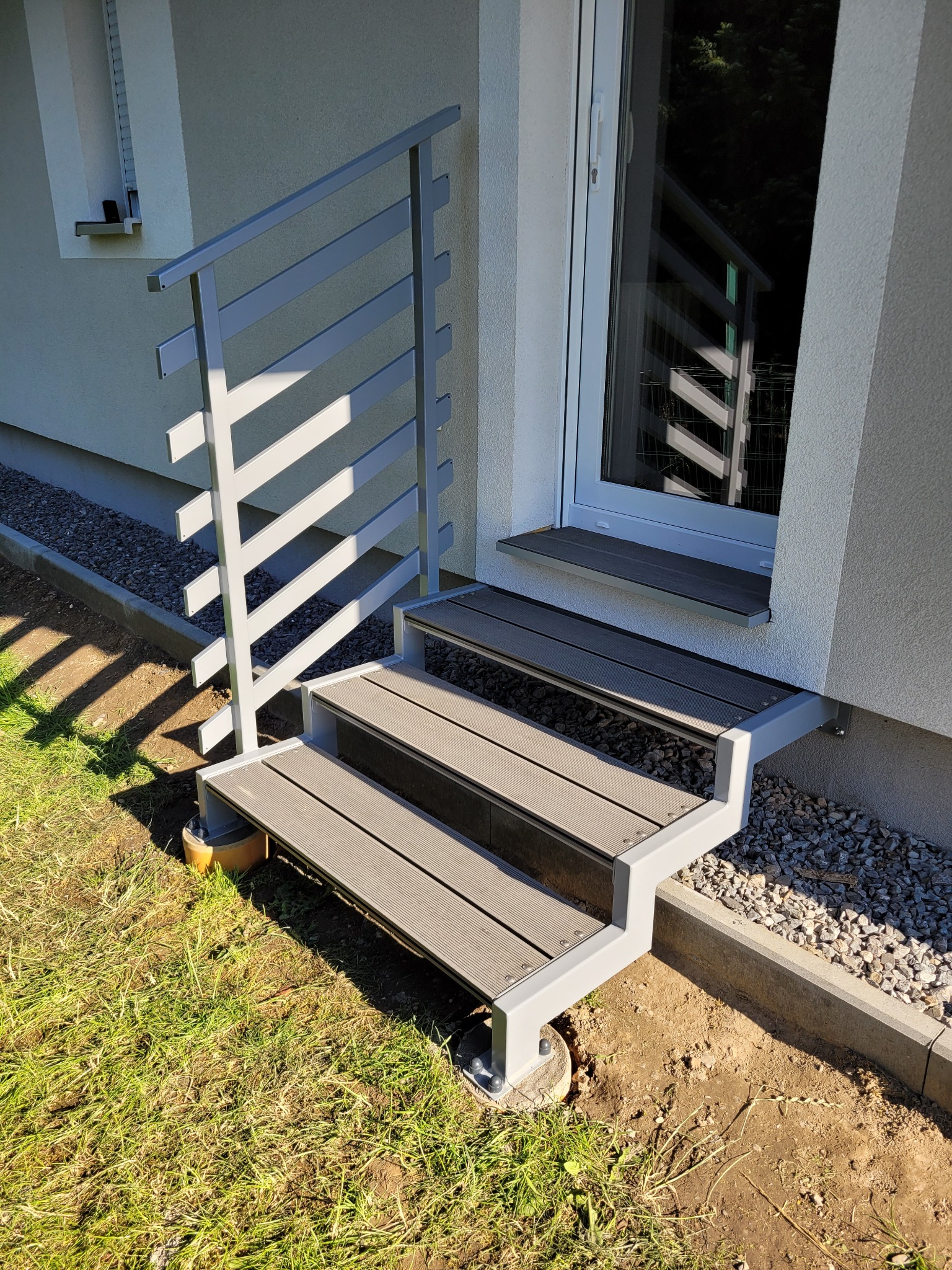 Zewnętrzne schody wejściowe z szarymi metalowymi poręczami i drewnopodobnymi stopniami prowadzące do drzwi balkonowych.