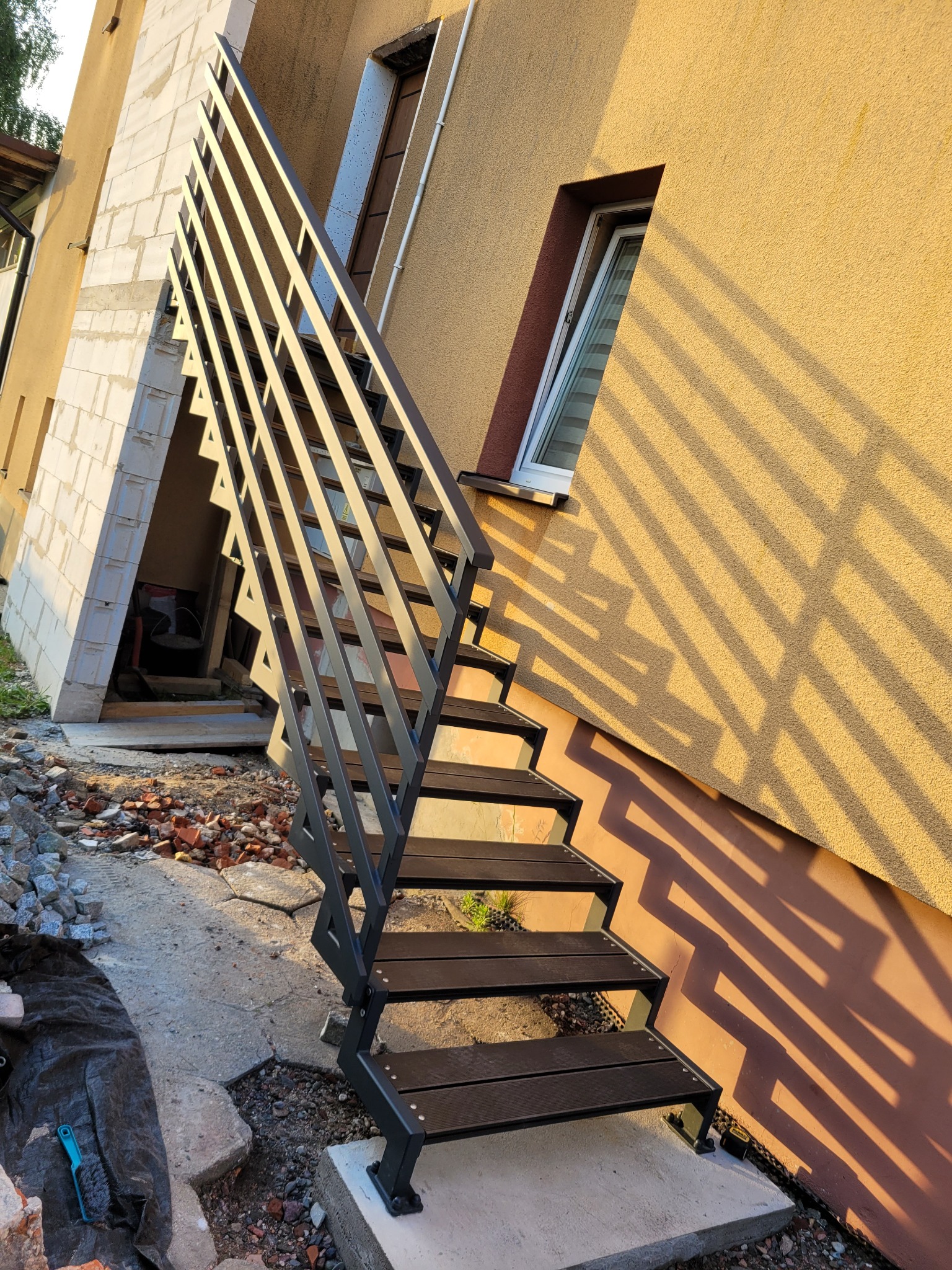 Zewnętrzne schody metalowe z drewnopodobnymi stopniami, prowadzące do wejścia w budynku o żółtej elewacji, częściowo w cieniu, z widocznymi elementami gruzu i przygotowania terenu.